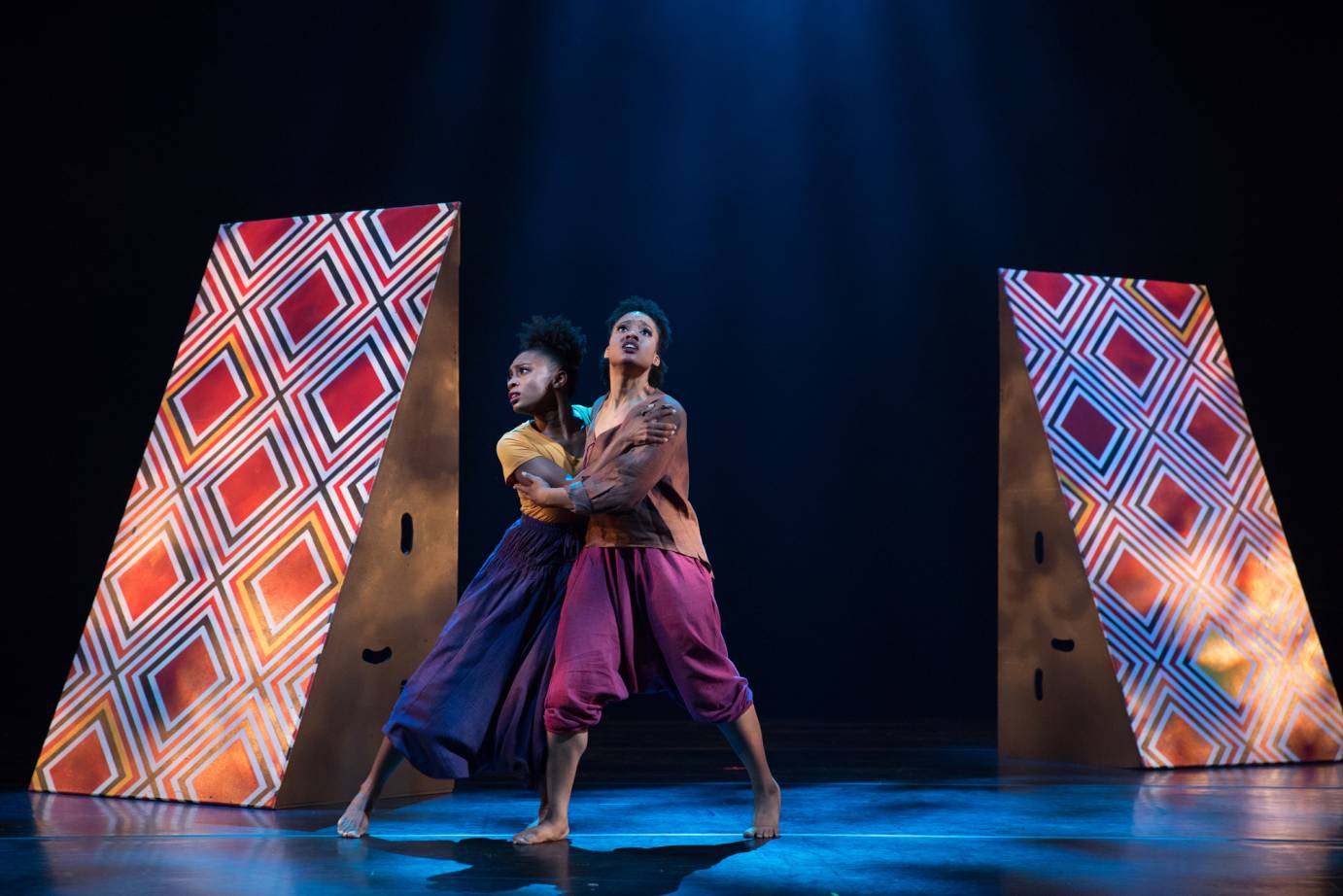 Two women clutch each other, looking fearful; two red-and-black folding props are behind them