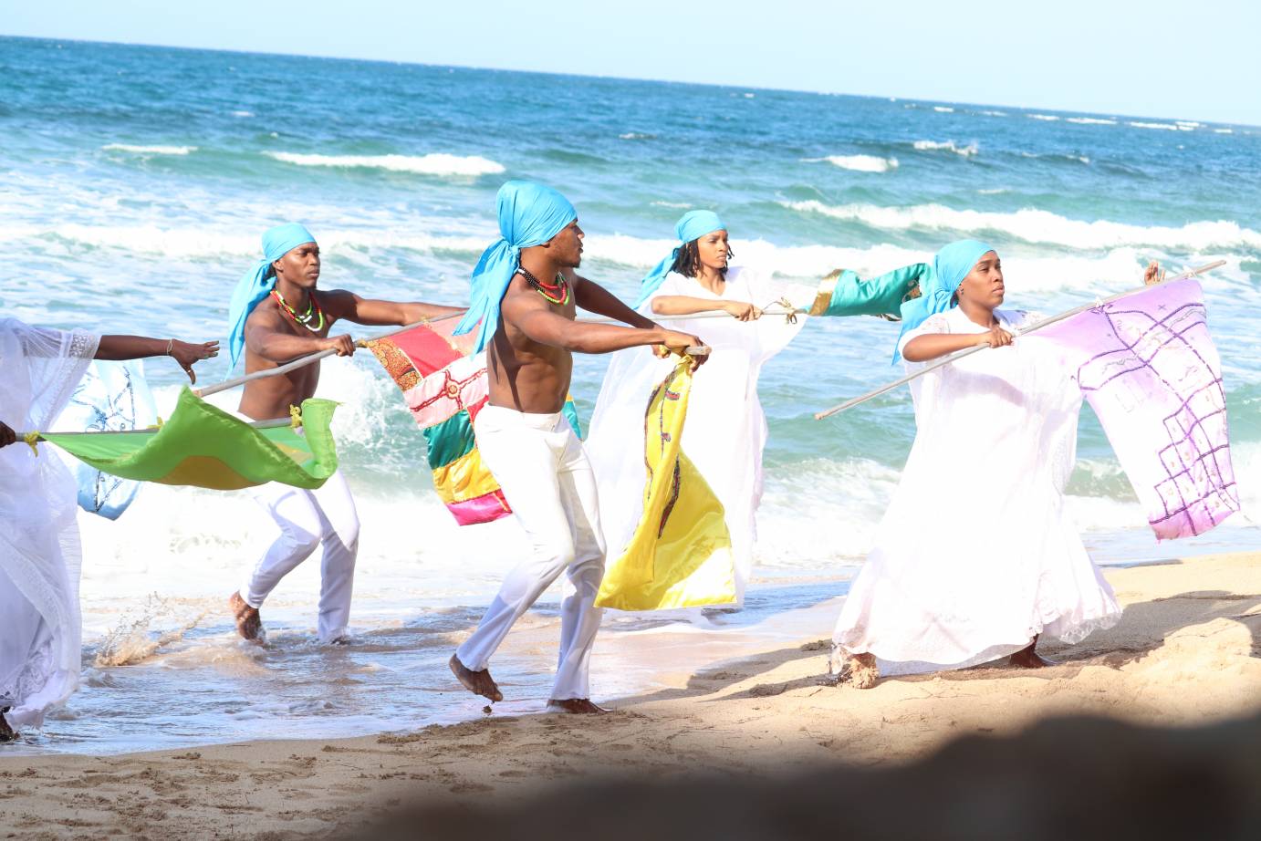 Against a turquoise ocean, men and women in white with aquamarine headscarves wave colorful flags