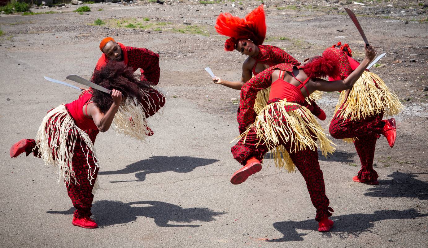 In a small circle, dancers in red with grass skirts lean sideways and lift one leg