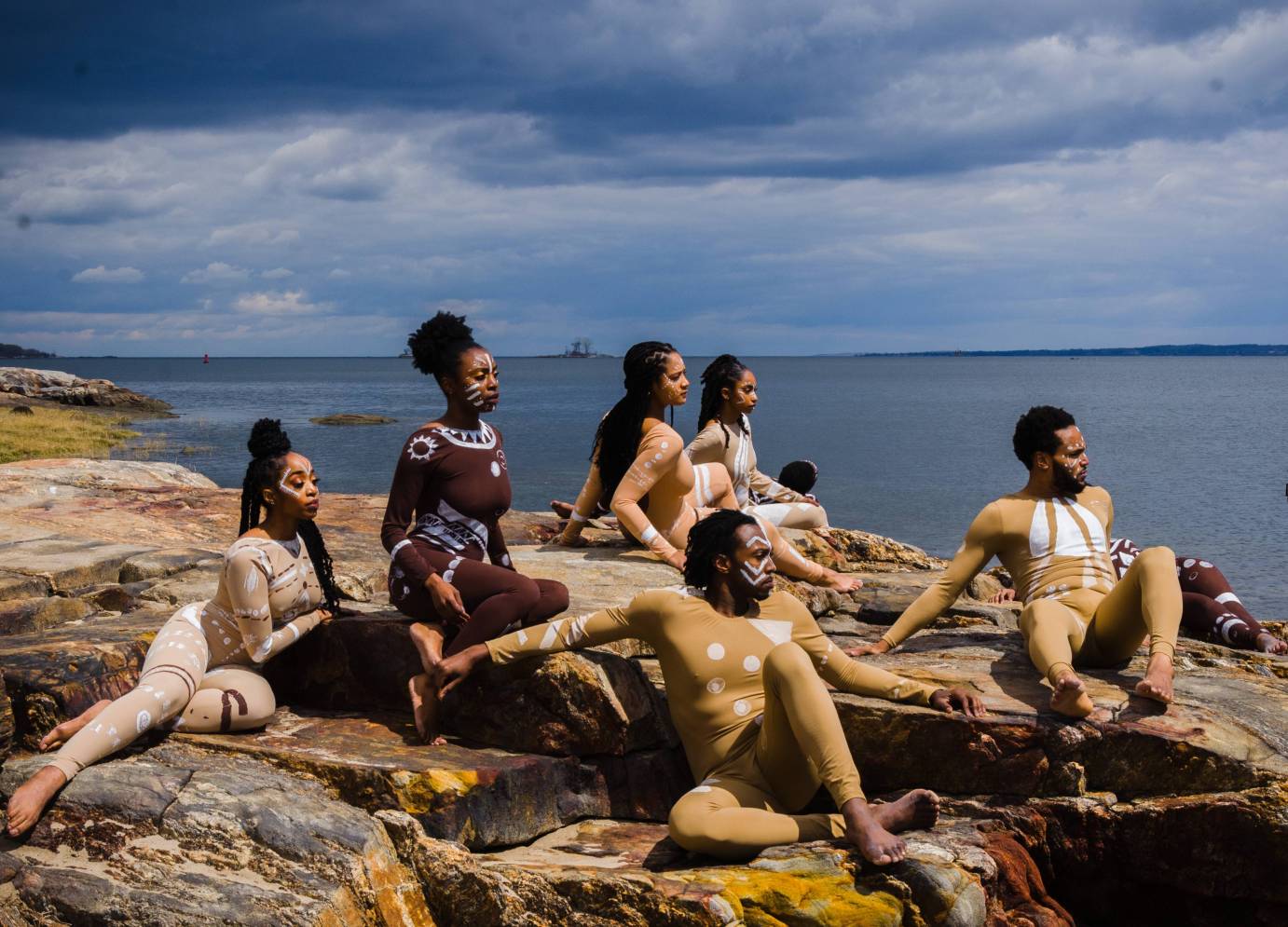 On a beach, mean and women in beige and brown costumes with white graphics lounge on rocks 