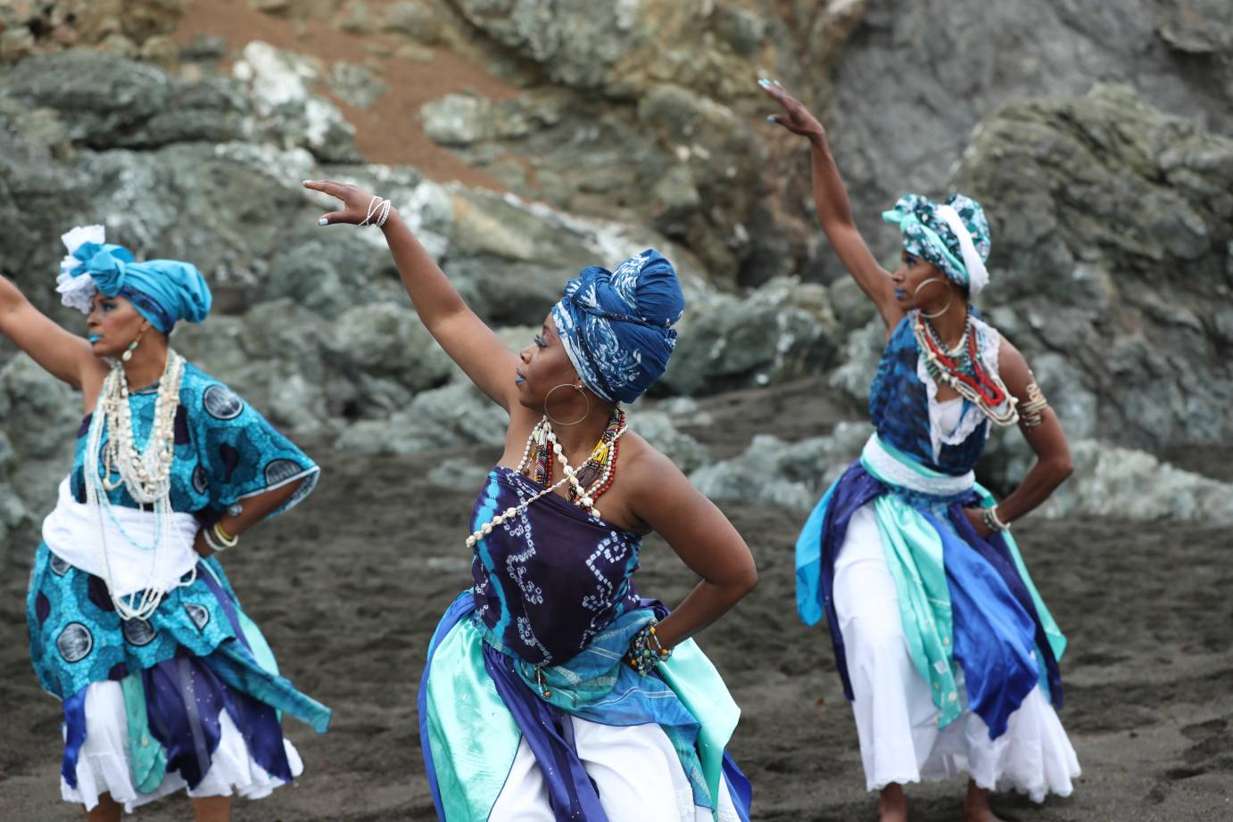 Three women in costumes of many shades of blue, lift one arm against a backdrop of rocks