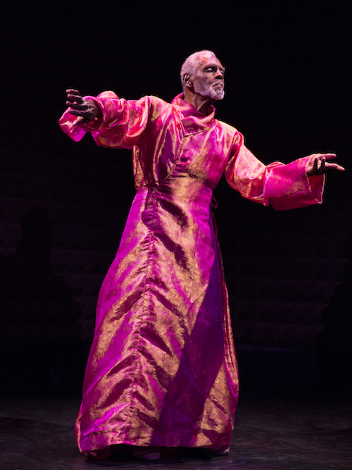 Gus Solomons jr wears a gauzy pinkish, orange robe. His arms are outstretched. One palm is turned up