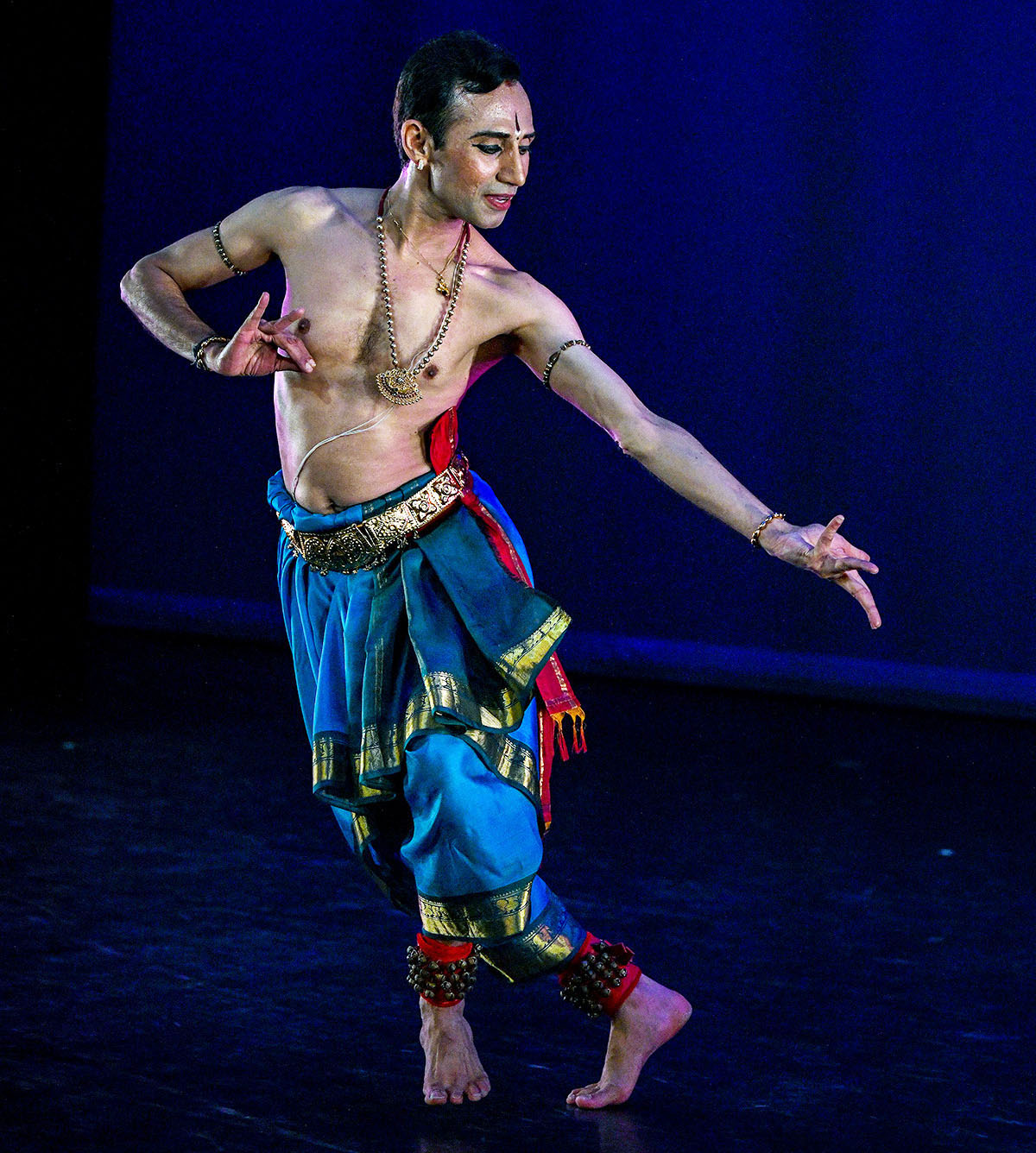 male Indian dancer strikes an asymmetrical pose... he wears blue baggy pants with gold trim and ankle bells around his legs