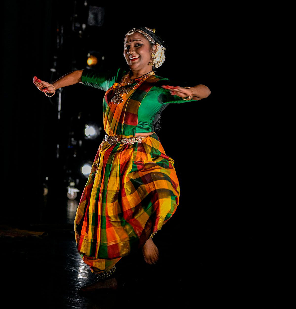 The woman Indian dancer jumps jubilantily. we notice not only her bejeweled hair but the rich green silk blouse and her madras checked skirt and scarf.