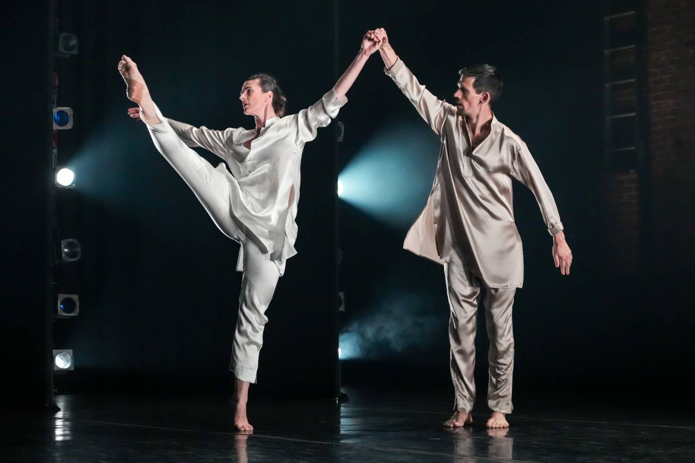 Woman and man dressed in silk beige longsleeved shirts and long pants. Hands held above their heads. Woman's leg extended to head. Heads each looking right.