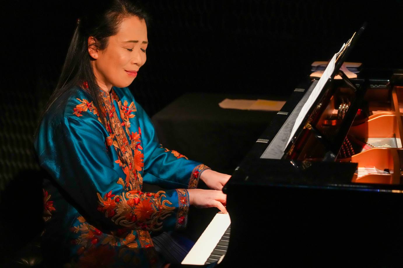 A woman pianist with long black hair looks down at the keyboard, a small smile on her lips.