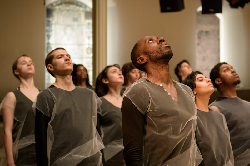 A group of people wear sheer netted tunics over black clothing. They stand en masse and look up to the ceiling.