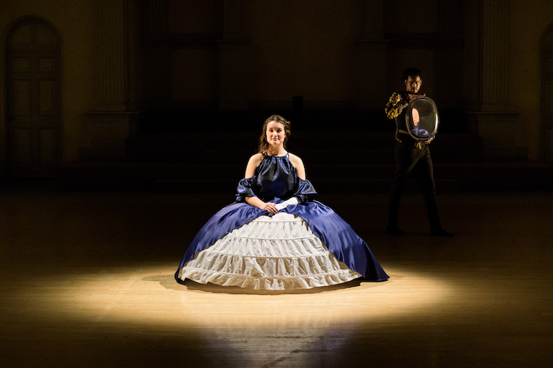 A woman in an old-fashioned ball gown and hoop skirt sits in a chair. A man holding a mirror looks at her in the background.