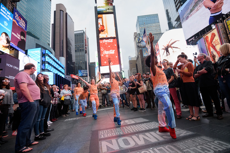 Dancers in red and blue costumes pointing their arms to the sky. A crowd, some people wearing 3-d glasses, stand around them.