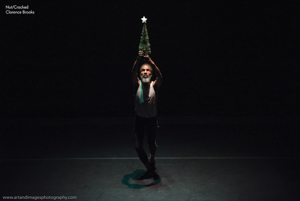 black man with grey dreads center stage holds a little Christmas tree high in the air