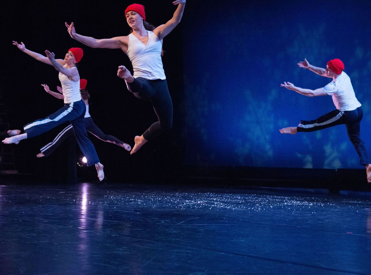 another jumping shot this time the dancers all aloft extend their arms in front of them and sport white t's, black striped track pants and bright red winter caps 