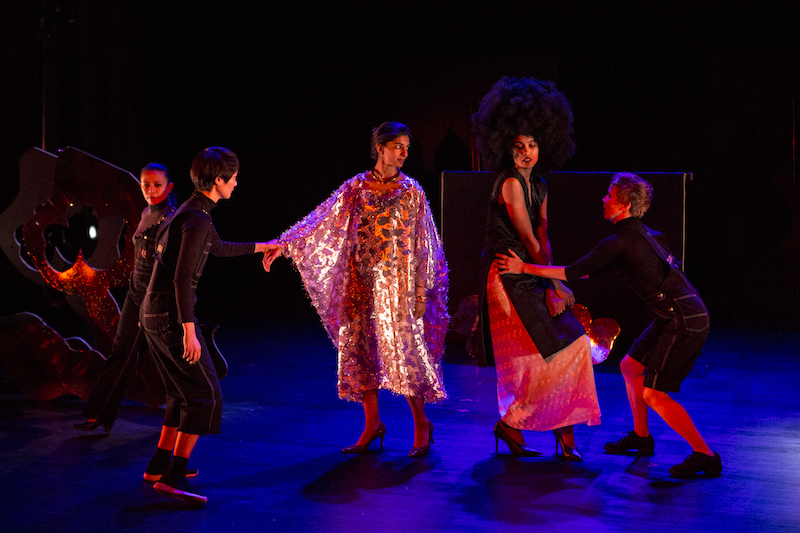 Two women in bedazzled, '70s inspired costumes are led by the hand by two performers in black overalls