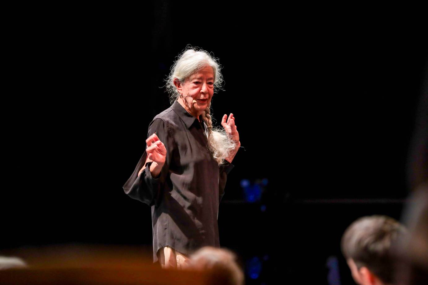 An elderly woman with white hair both hands raised chest high, addresses the audience seated closeby.
