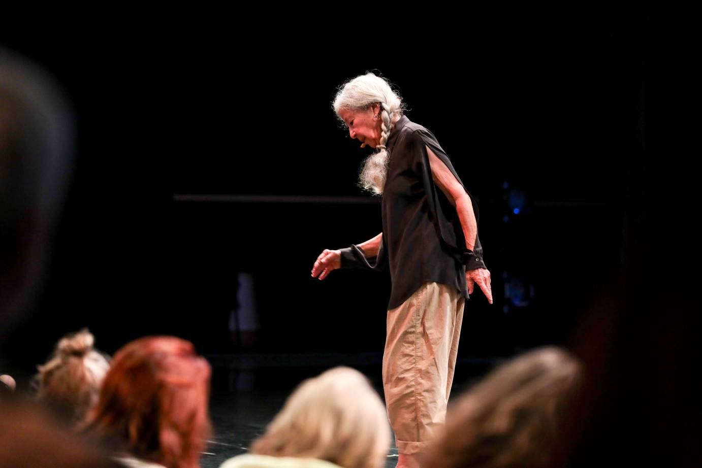 An elderly woman with chin lowered, white braid hugging her face and reaching down the front of the left side of her chest, pauses in a brown blouse with a slit up the arm.