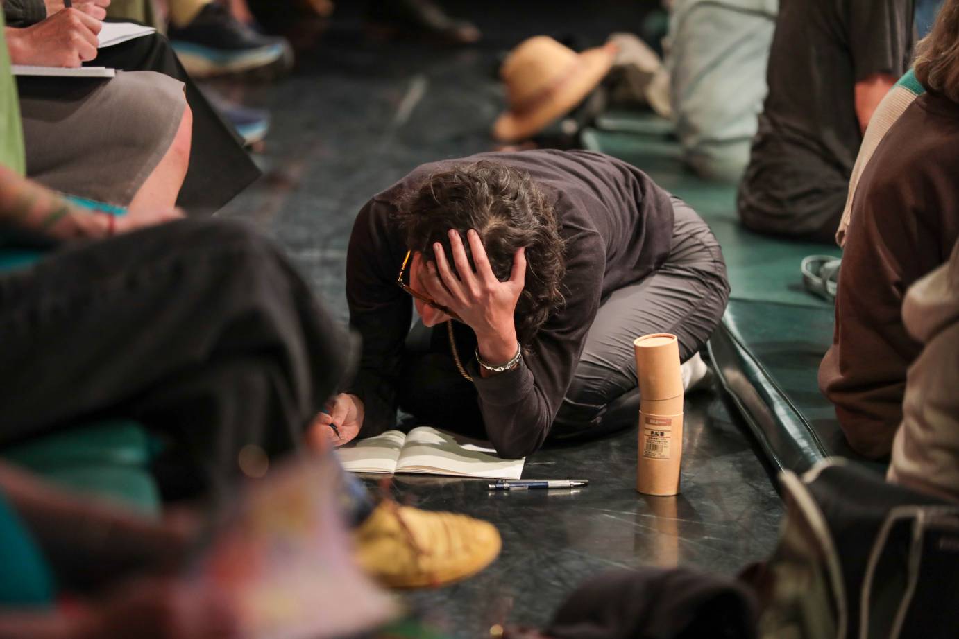 An audience member is crouched on the floor with one hand covering their head concentrating on their writing.