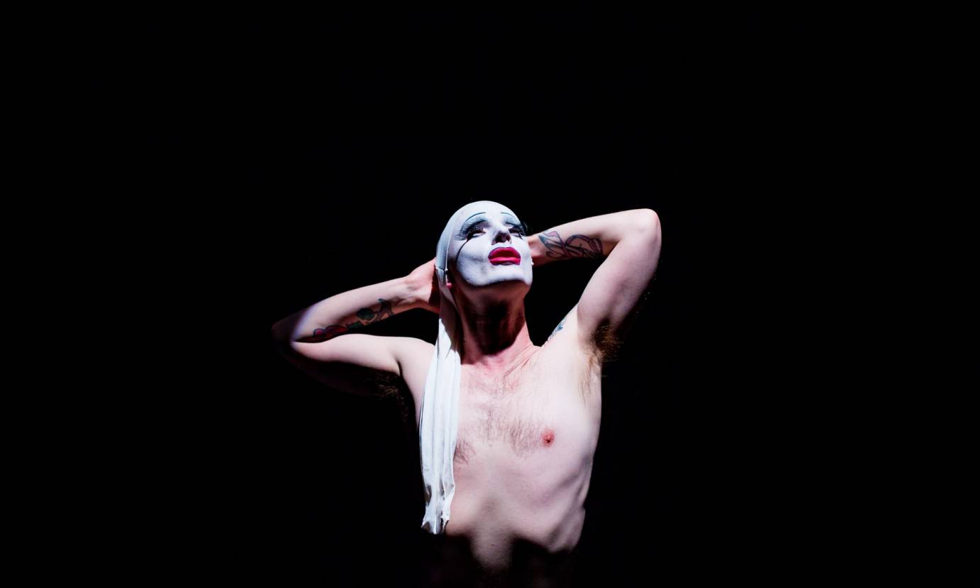 Dickie Beau, shirtless, places his tattoo-ed arm behind his head. His face, painted white with bright red lips, looks up to the stars.