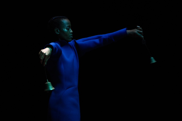 in profile Dorothee Munyaneza stands against a dark background, her cheekboneis highlighted by glitter, her eyes closed. She holds waves her arms in opposite directions as rings bells