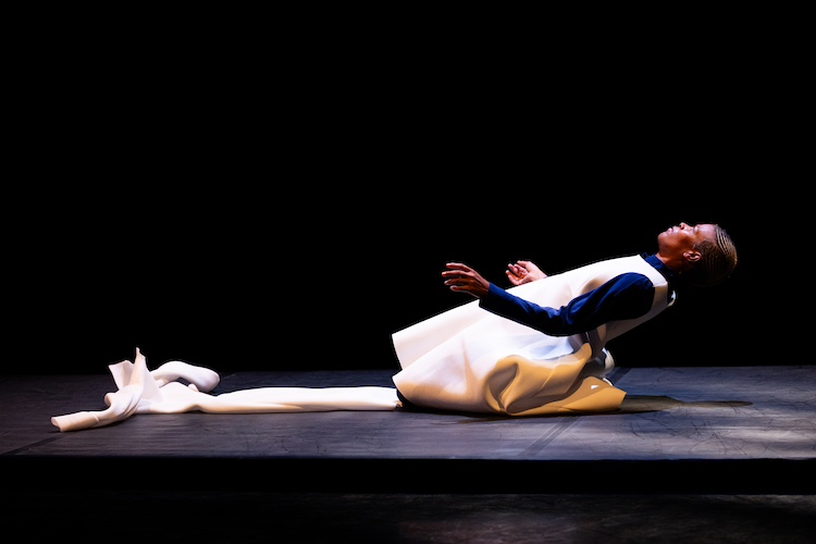 artist Ife Day, on her knees, leans backward, creating a long line. Over her blue outfit, she is wearing what seems to be part of a straight jacket . across from her another straight jacket or what seems to be one lays on the floor.