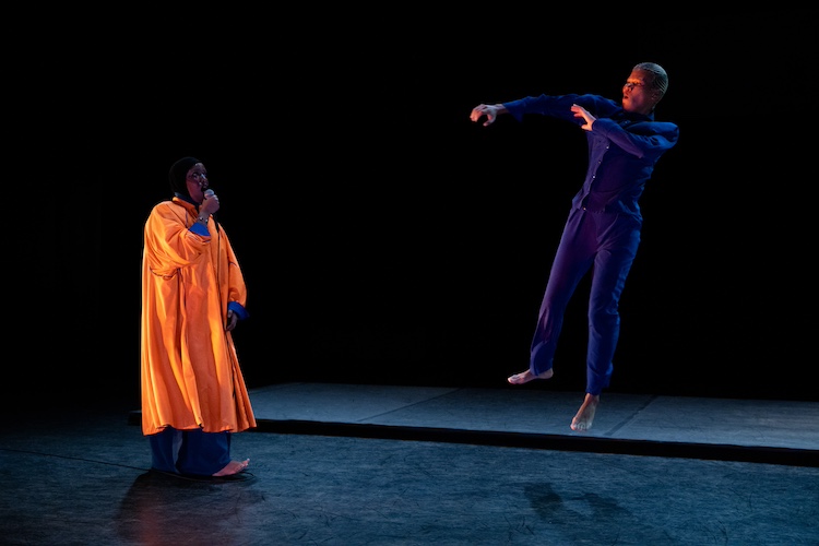 Asmaa Jama donned in a large robe of orange over a blue gown sings to the artist Ife Day, directly across from her. Ife sails vertically aloft her arms in front of her with an expression of joy on her face. She is wearing a blue jumpsuit.