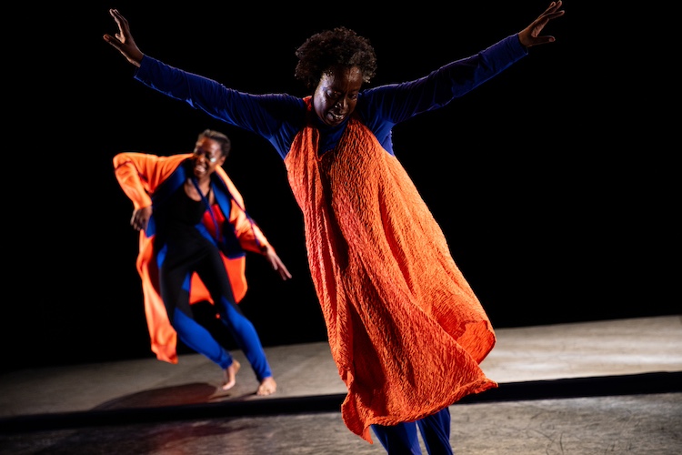 two dancers, in blue and orange, in front Nido Uwera her arms extended in a V upward twists, gazing at her feet. Behind her Dorothee Munyaneza appears to be mid-skip.
