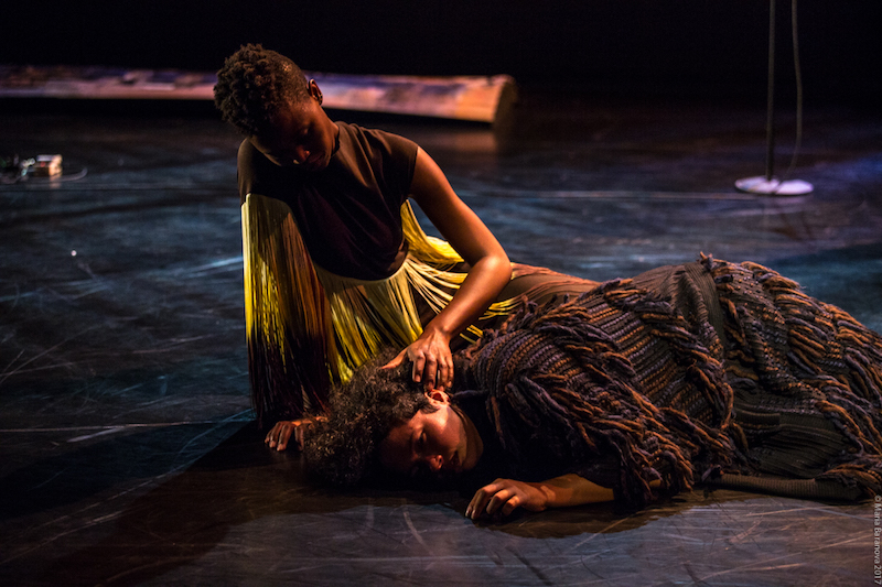 Two women sit on the floor. The seated women strokes the hair of the other who is laying on the ground.