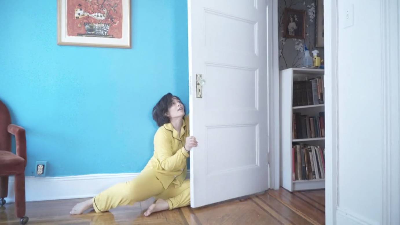 A woman dressed in yellow hugs a door against a turquoise-painted wall