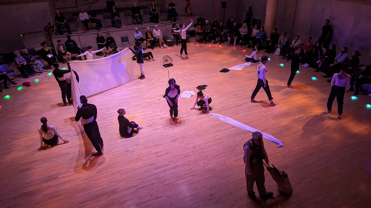 The theater-in-the-round seen from above with audience seated in chairs and colored-gel hand sculptures placed in a circle in front of them.. The stage floor, a pink glow, is scattered with dancers performing seated and stationary, standing and reaching legs wide apart, and holding up sheets.