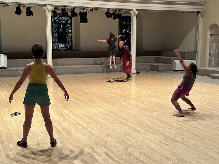 dancers in colorful rehearsal clothes spread around the dance floor, one has her back to us, another in profile seems in midst of a jig, in the background a dancer is caught in a jump, while a musican plays a keyboard on the floor with his toes