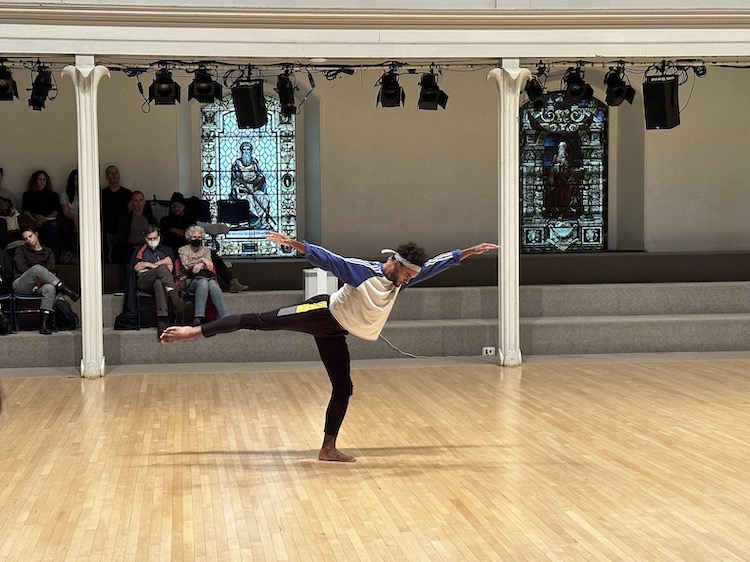 NYCBallet principal dancer Taylor Stanley in an expansive arabesque. They are  wearing sweats, a baseball shirt , and bandana and is sporting bare feet