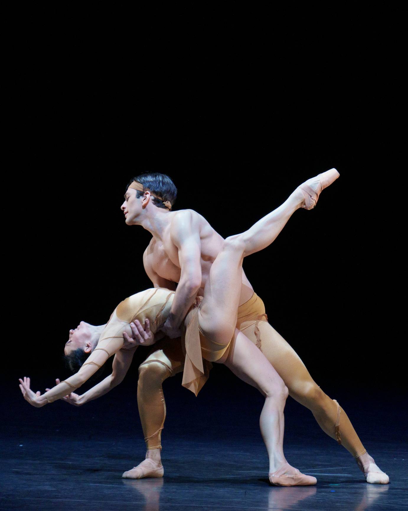 A ballerina in a crab-like position, falls backward with an extended leg in attitude and both arms curved behind her head. She is held at the waist by her standing partner.