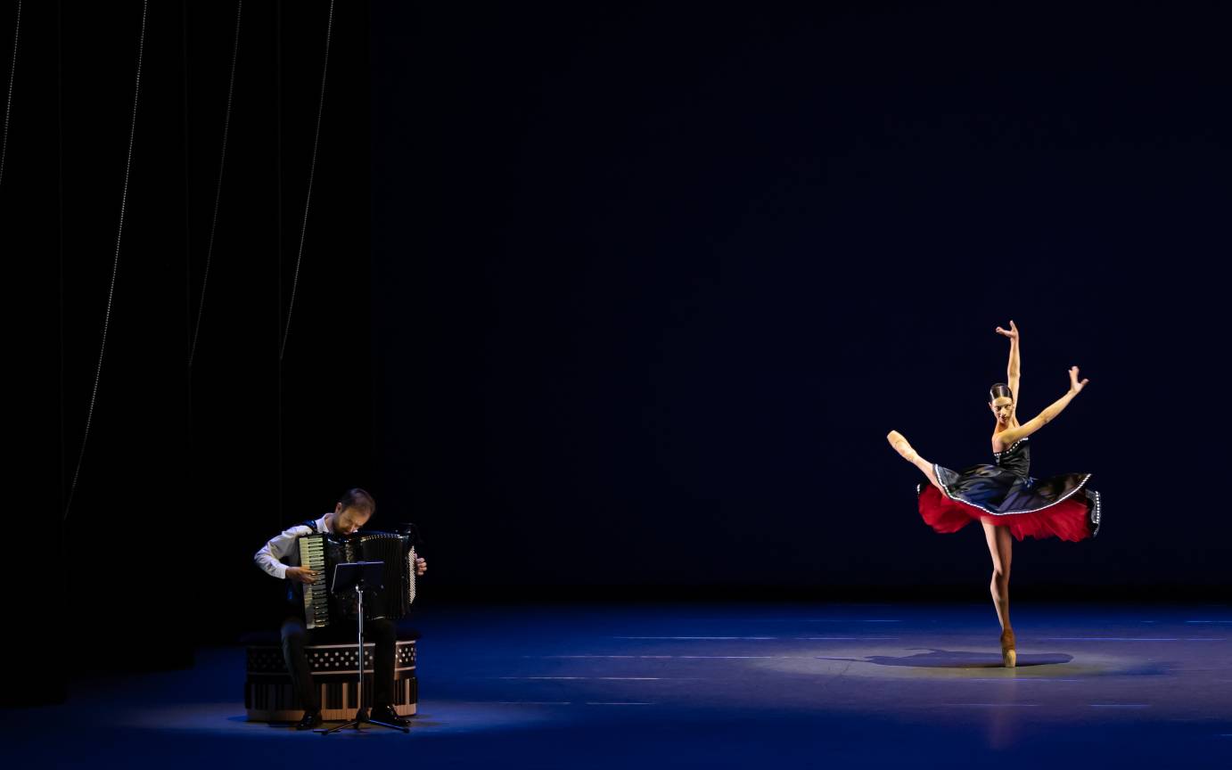 A woman in a sleeveless black costume lined with red exuberantly lifts one leg and her arms. She is accompanied by a seated accordianist. 