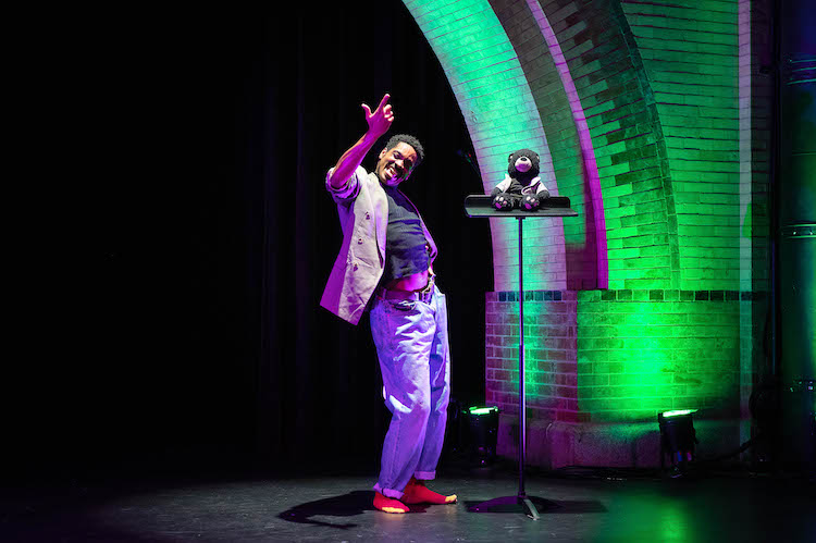 under the archway of emoves stage Ja young black man in jeans and a tan sport coat is engaged in a vibrant conversation with a black teddy bear doll who sits on a music stand