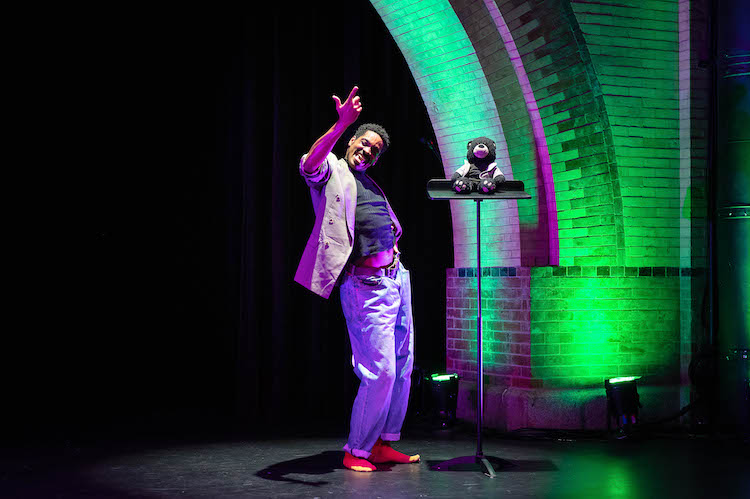 under the archway of emoves stage Ja young black man in jeans and a tan sport coat is engaged in a vibrant conversation with a black teddy bear doll who sits on a music stand 