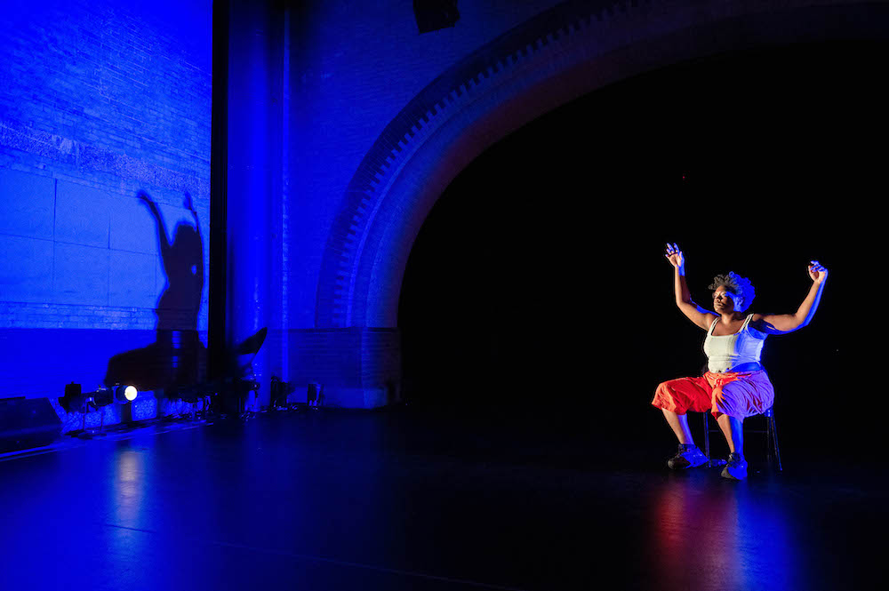 Ogemdi Udi a Black woman in a white tank top, orange pants, and sneakers sits on a chair arms raised to resmembe a goal post. We notice the dark blue shadow her form creates against the wall of Harlem Stage.