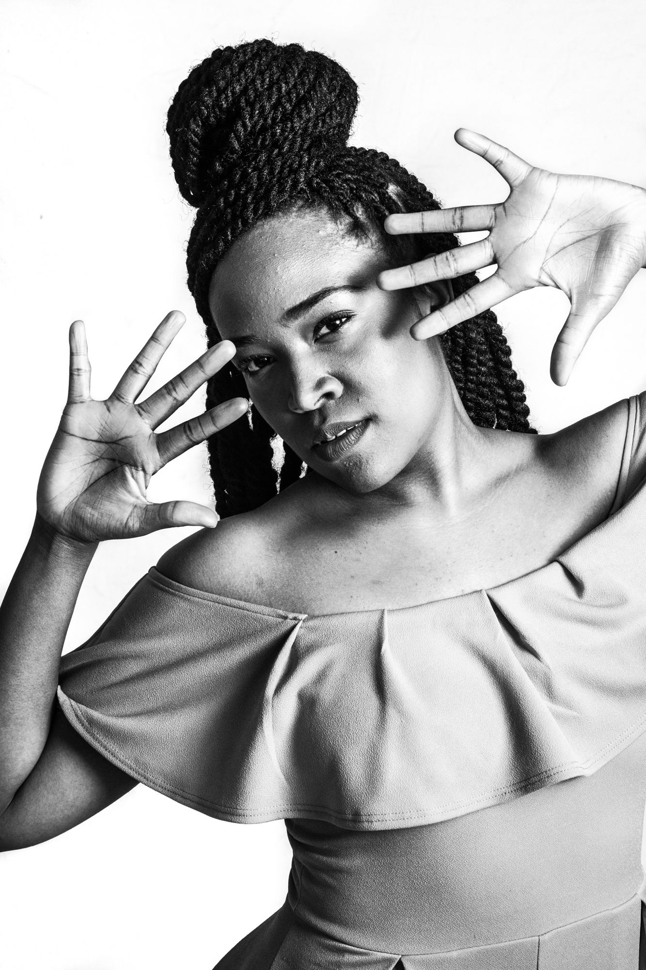 a black and white portrait of dancer Brittany Stewart. Her torso curves to the right, as she looks out at us through spread fingers that cover the side of her face. Her hair is braided with a braided bun, like a crown at the top of her head with the rest of the braids cascading down her back. She is wearing an off the shoulder leotard  with one large ruffle at its top 