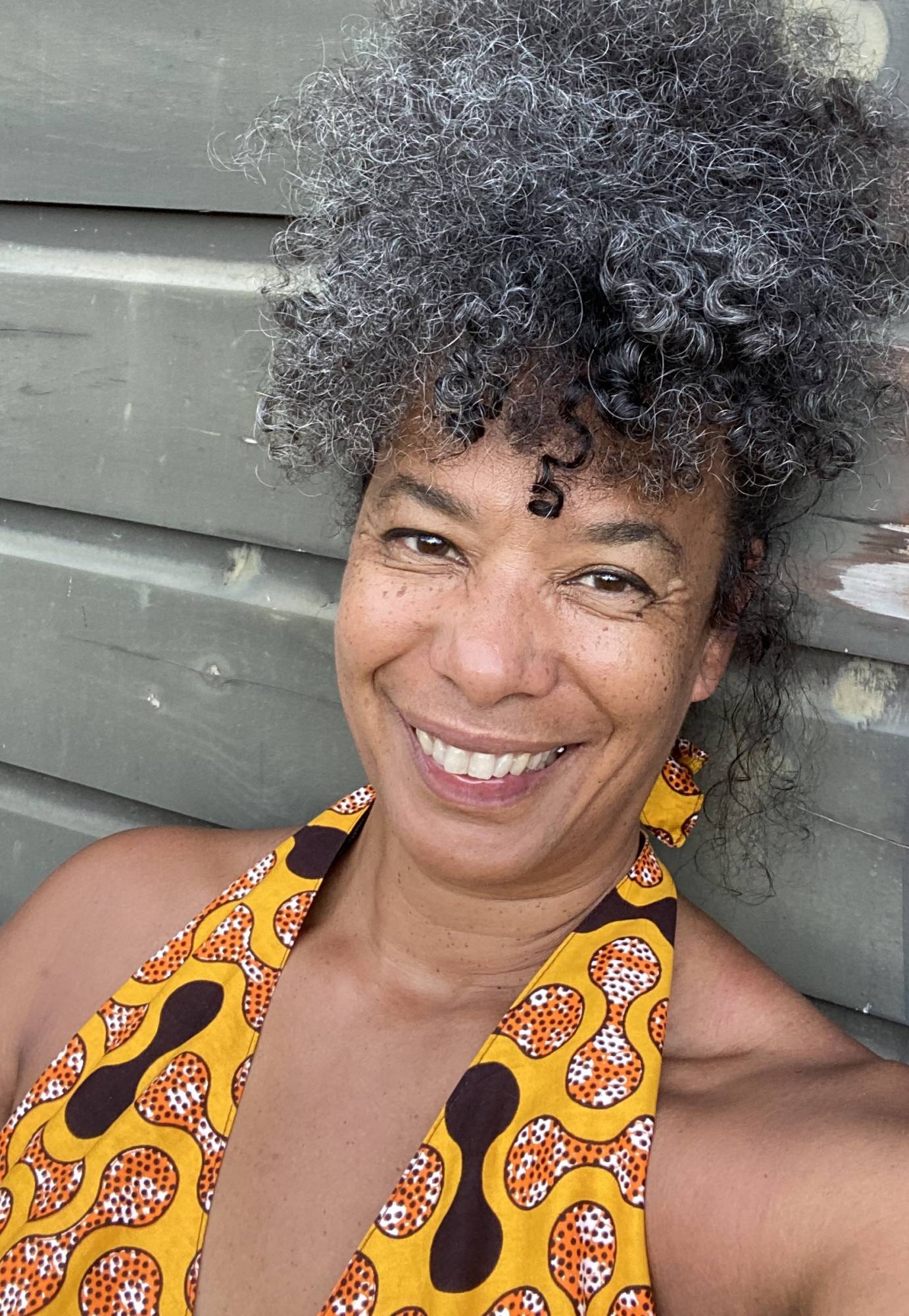 Edissa Weeks, a light skinned Black woman with greying curly hair tied up in a bun, smiles beautifully, in her halter sun dress with an African print of yellow and orange. She leans against a grey wall. 