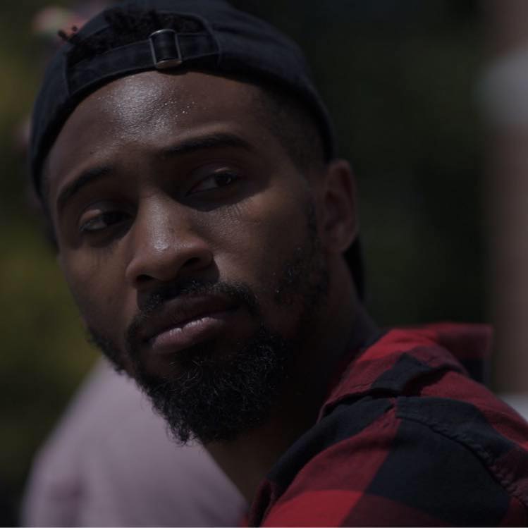 The artist Johnnie Cruise Mercer's head shot. They, a black male appearing person with a mustache and beard peers sideways as if someone called his name. He wears a black baseball cap turned backwards on their head and they appear to be wearing a red and black checked flannel shirt