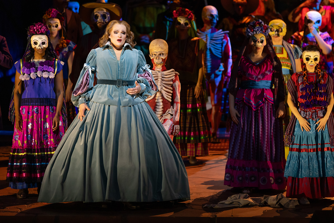Greta Garbo, in grey blue long skirt, stands before the group of skeletons and masked townspeople.