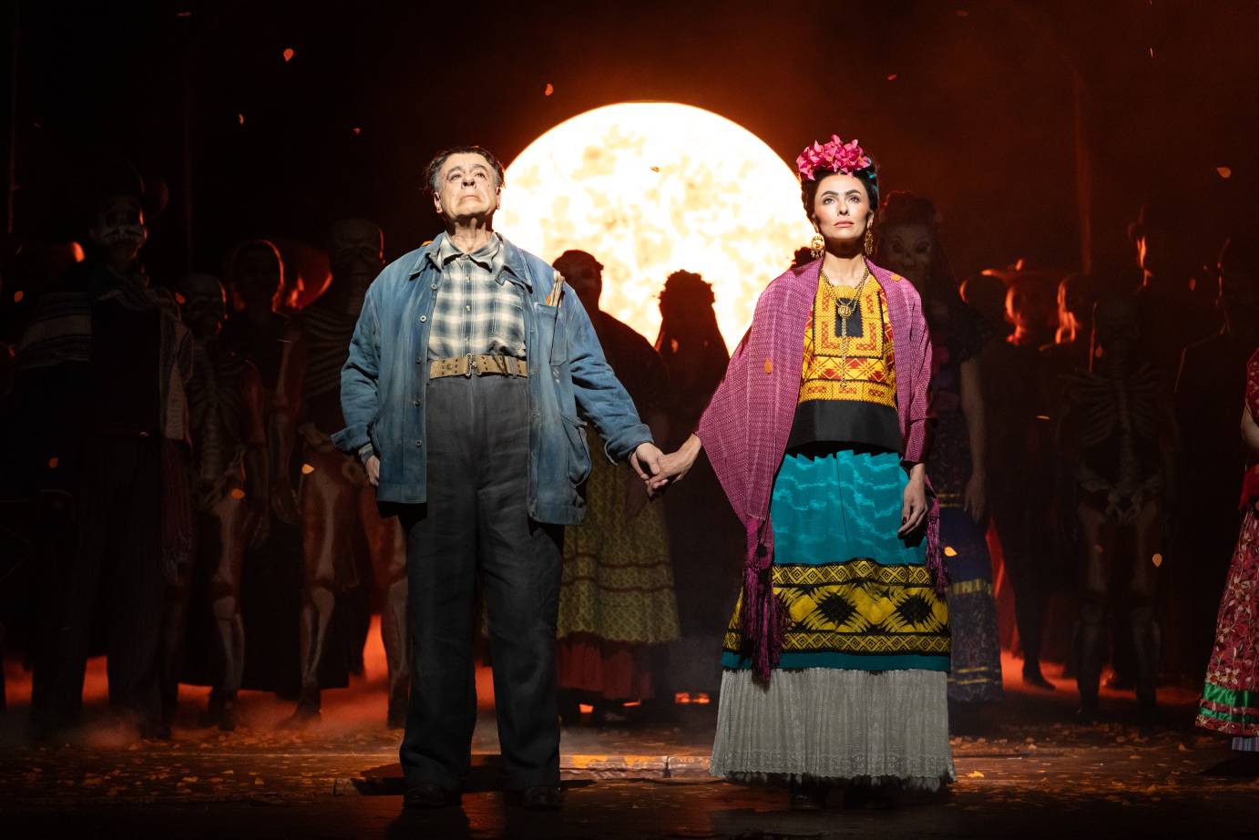 Diego, dress in relaxed blue pants and jacket, holds hands with Frida, costumed in colorful top, long skirt, and shawl with pink flowers atop her head. They stand looking forward their shadows projected onto a moon behind them.