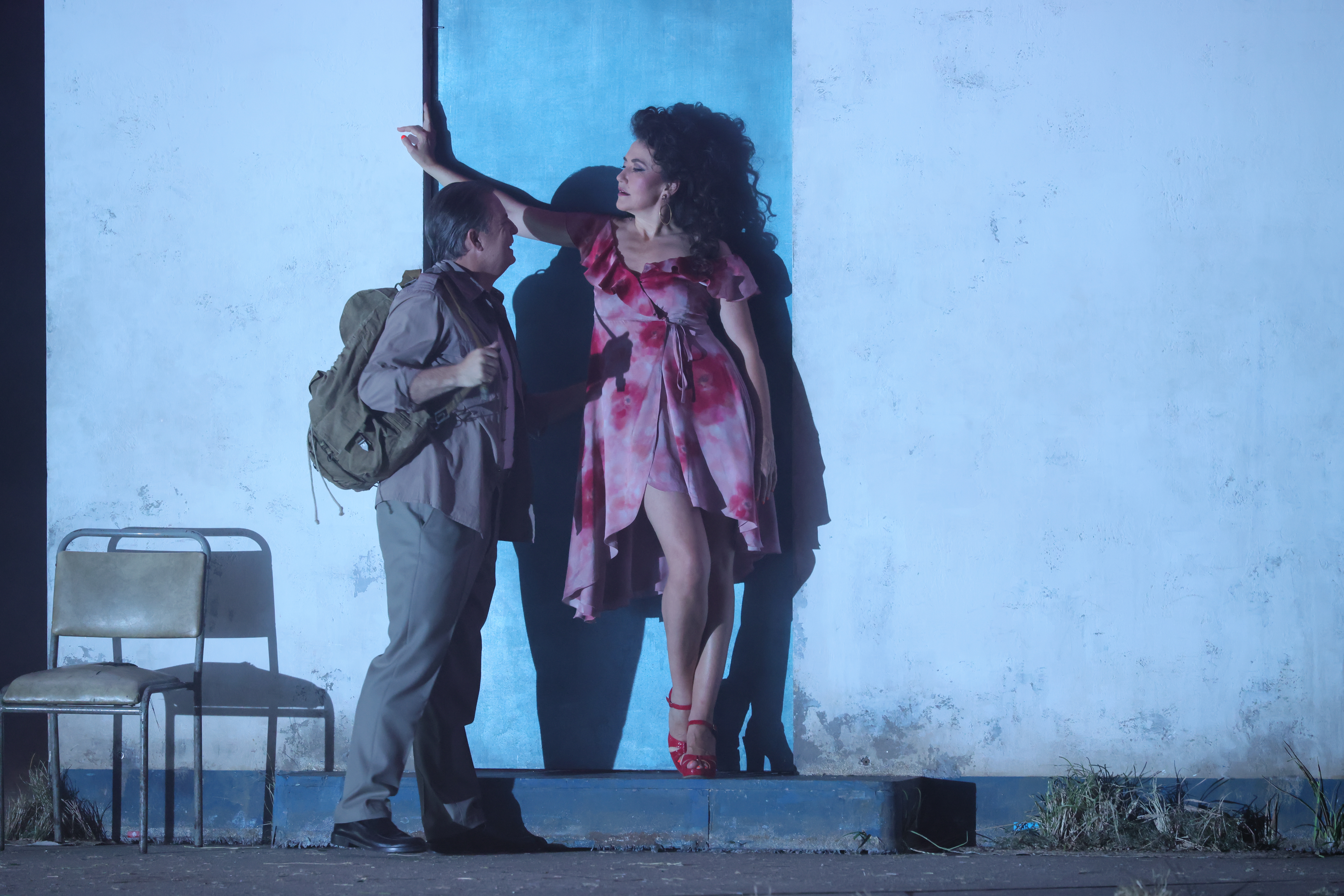 Carmen, in pink flowered dress, leans against a blue wall looking down on Don José who stands close to her.