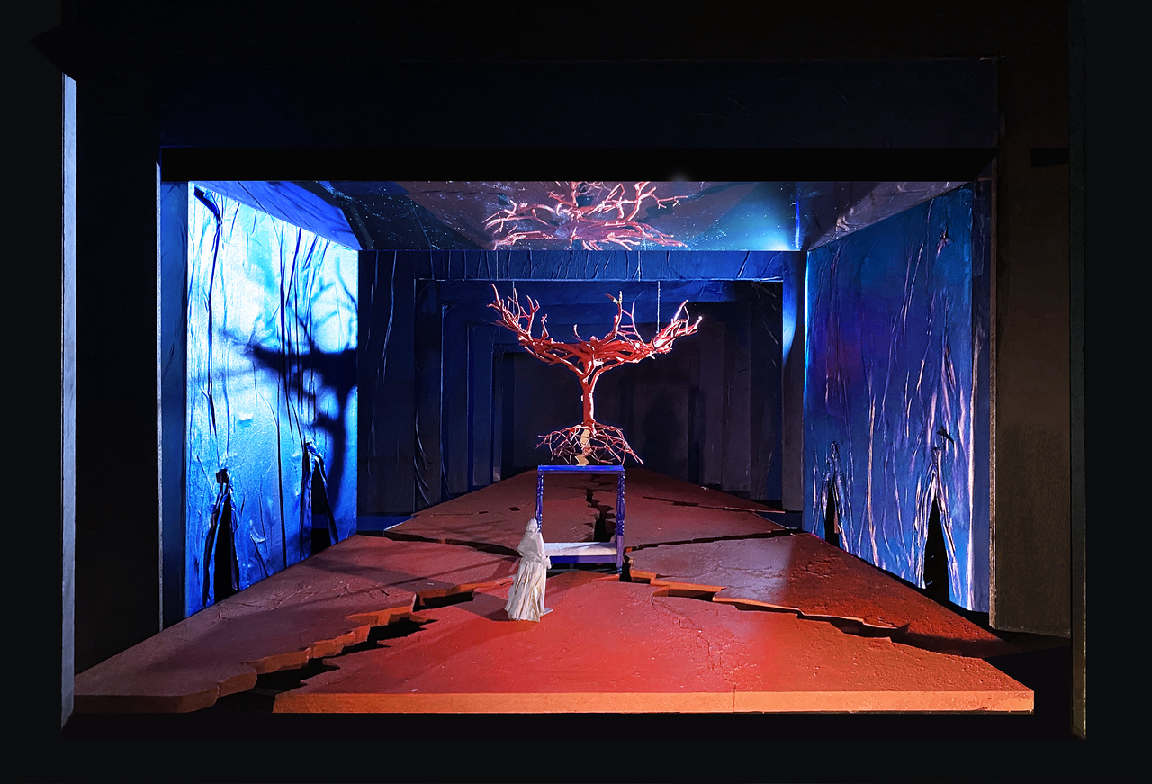 The stage set of the Met: a figure in white stands near a susupended tree reflected in a mirror above. The red floor is marked by jagged openings. The sides are a 