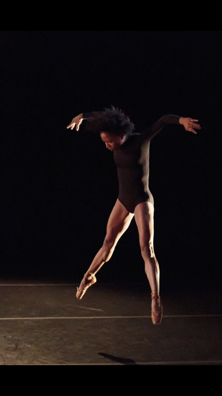 A girl in pointe shoes jumps in the air, shoulders rolled forward, arms curved downward, one leg extended slightly