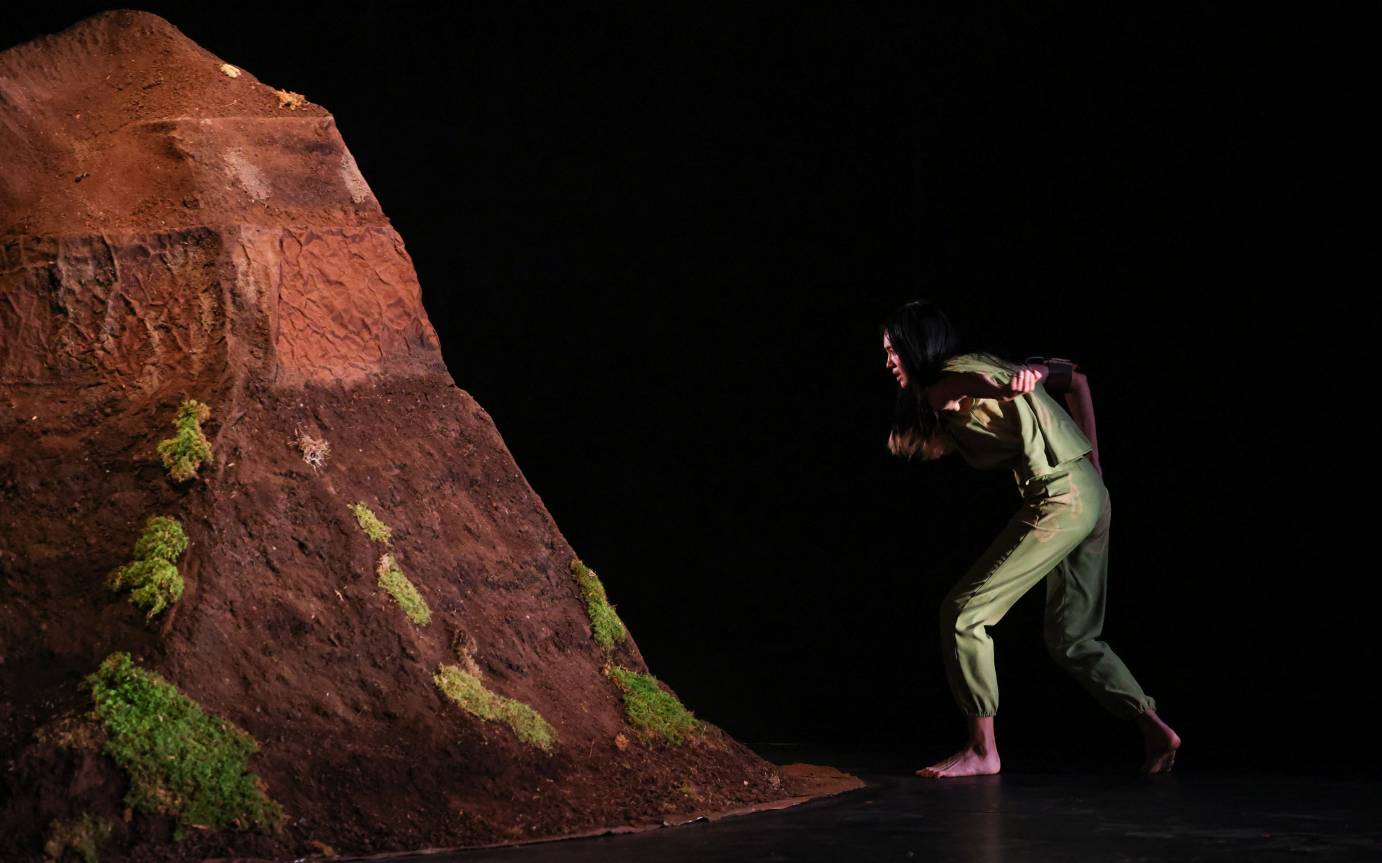 a mound of dirt piled high on stage left, a dancer in green peers at it
