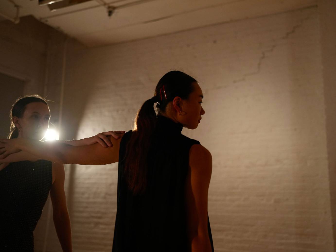 One dancer standing ahead of the other, her head in profile as a white light shines between them..