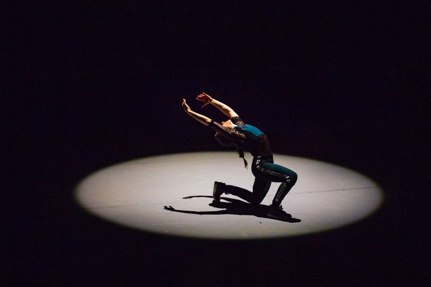 Ephrat Asherie kneels in a spotlight, her arms lifted above her head