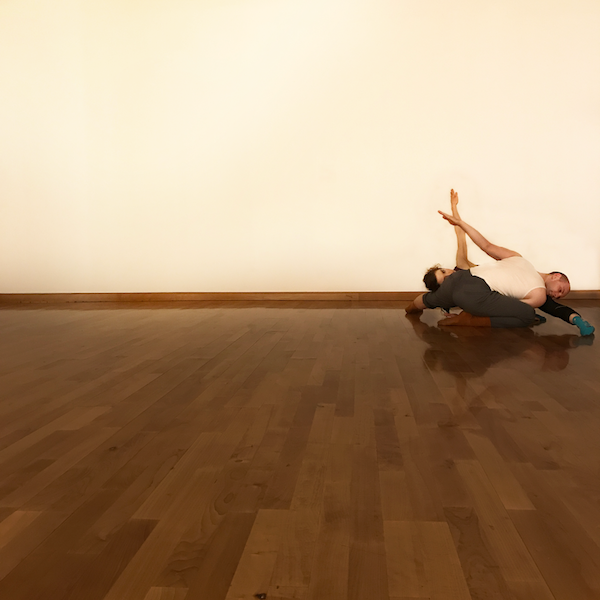 Two dancers are laying on the floor in rehearsal clothes. Their bodies fold over one another.