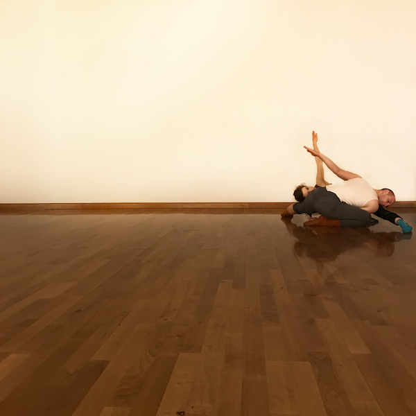 Two dancers are laying on the floor in rehearsal clothes. Their bodies fold over one another. 