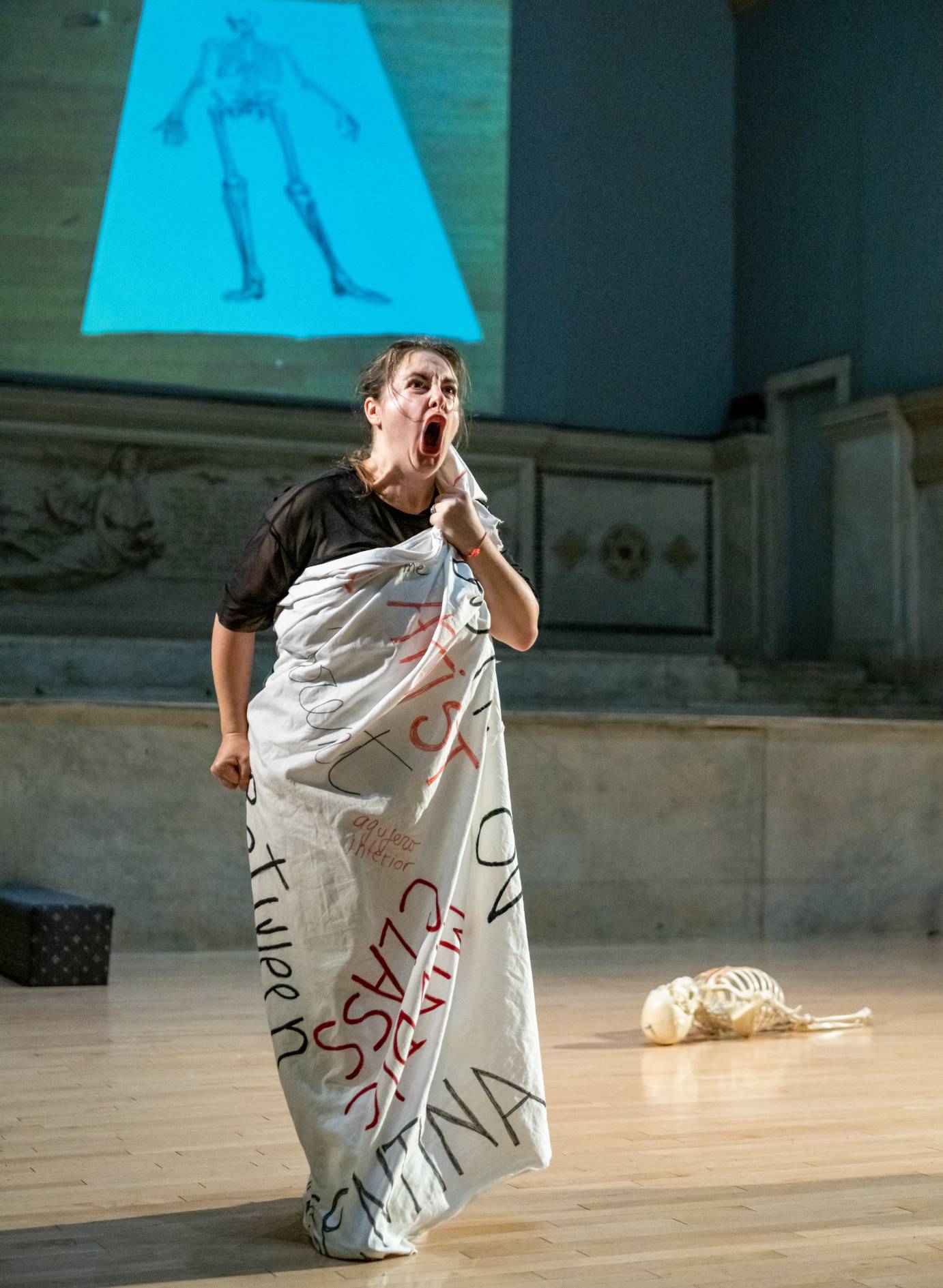 Latina woman facing the audience dressed in a white shroud that bear the words, CLASS, ARTIST, etc. The woman's fists are clenched and she is angrily shouting.