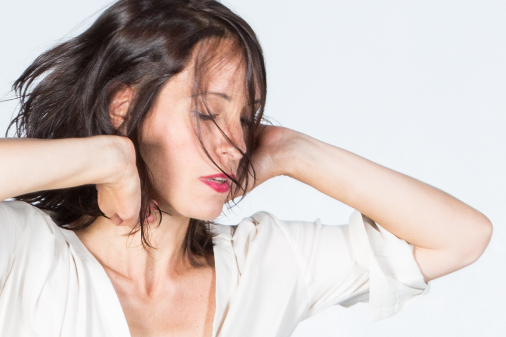 White woman with soft brown, chin length hair, in a 3/4 profile, casting eyes downward, with hands caressing her neck, dressed in a white blouse