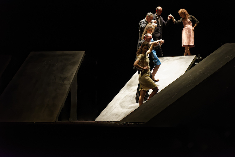 Dancers stand on ramps that are at various levels on stage. They hold hands doing a grapvine motion.